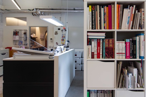 In the foreground a bookshelf, in the background plan cabinets with a standing workstation and cutting tools